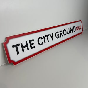 Op de afbeelding: Wit straatnaambord met rode rand en zwarte tekst die "THE CITY GROUND NG2" luidt.