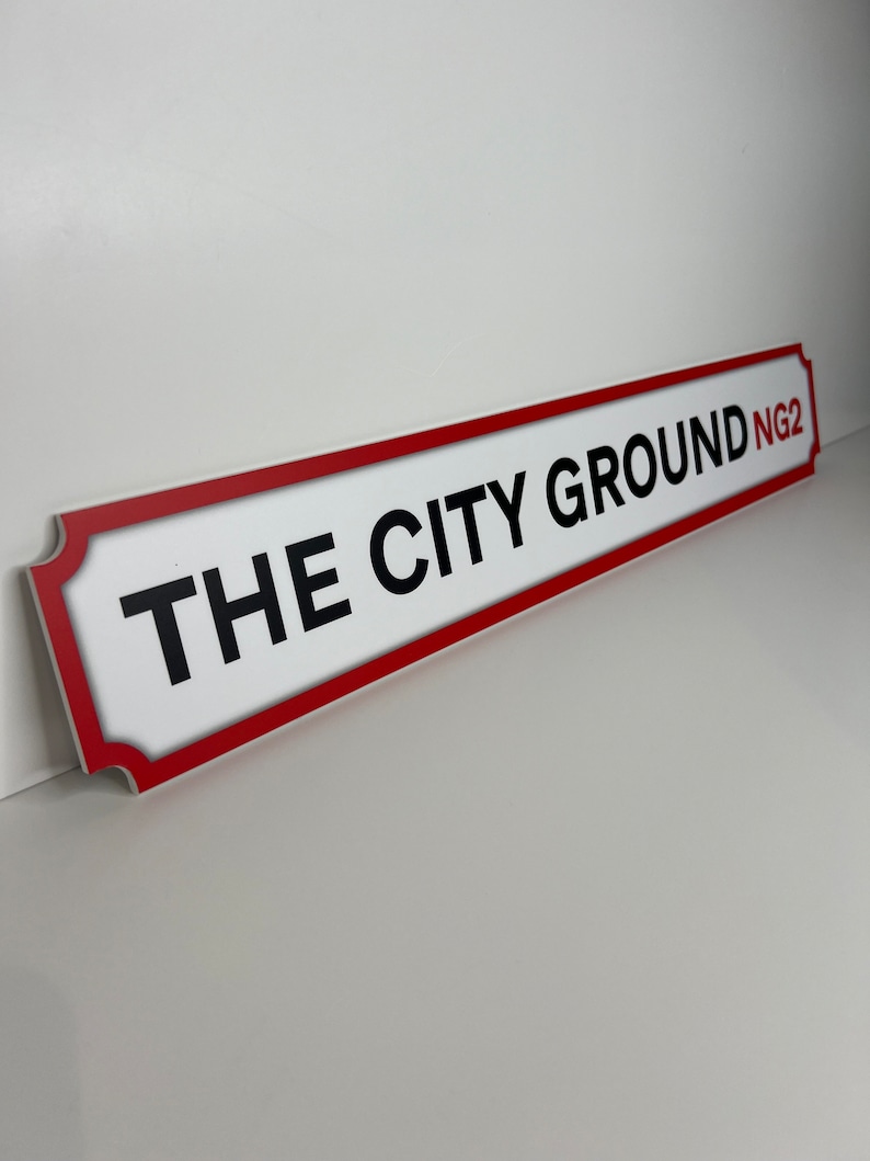 Op de afbeelding: Wit straatnaambord met rode rand en zwarte tekst die "THE CITY GROUND NG2" luidt.