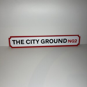 Op de afbeelding: Een rood en wit bord met afgeronde hoeken en zwarte tekst die "THE CITY GROUND NG2" luidt.