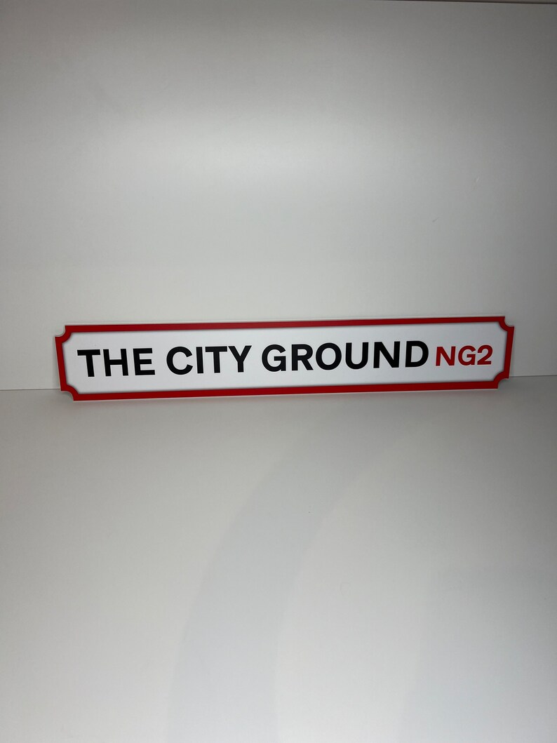 Op de afbeelding: Een rood en wit bord met afgeronde hoeken en zwarte tekst die "THE CITY GROUND NG2" luidt.