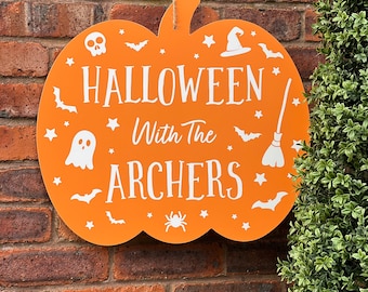 Pode incluir: Um letreiro de madeira laranja em forma de abóbora com o texto "Halloween With The Archers" em branco. O letreiro é decorado com morcegos brancos, estrelas, um fantasma, uma aranha, um chapéu de bruxa e uma vassoura.
