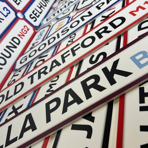 Op de afbeelding: Een verzameling witte en rode straatnaamborden met zwarte letters. De borden zijn op elkaar gestapeld en bevatten de namen van beroemde voetbalstadions, zoals Old Trafford, Goodison Park en Emirates Stadium.