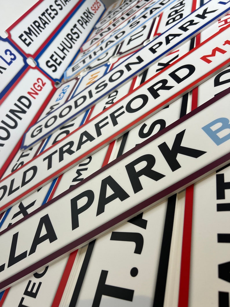 Op de afbeelding: Een verzameling witte en rode straatnaamborden met zwarte letters. De borden zijn op elkaar gestapeld en bevatten de namen van beroemde voetbalstadions, zoals Old Trafford, Goodison Park en Emirates Stadium.