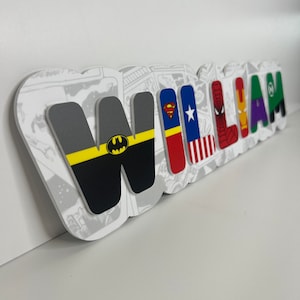 May include: A white background with a name spelled out in colourful letters. Each letter has a different superhero logo on it. The letters spell out the name "WILLIAM".