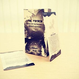 Könnte beinhalten: Ein Kartenspiel-Box und Karten mit dem Titel "THE PATRON DECK OF PATRONS QUEST-GIVERS". Die Box hat eine Schwarz-Weiß-Grafik. Zwei Karten werden angezeigt, eine flach und eine aufrecht. Die Artikel befinden sich auf einer hellfarbigen Holzoberfläche.
