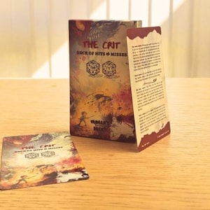 May include: A tabletop game box and card set titled "THE CRIT Deck of Hits & Misses." The box and card feature a fantasy-themed illustration with dice and text. The box is open, revealing a card and an insert.