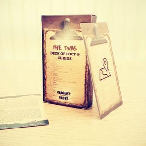 May include: A vintage-style box and cards for a game called "The Swag Deck of Loot & Curios." The box and cards have a weathered, antique appearance with text and a map icon. The cards are on a light-coloured surface.