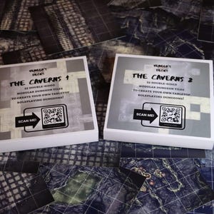Puede incluir: Dos cajas blancas, "The Caverns 1" y "The Caverns 2", para juegos de rol de mesa, con texto y códigos QR. Están sobre una superficie con un diseño de mapa de mazmorra.
