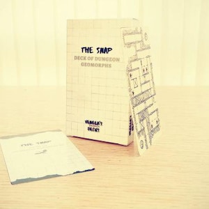 Peut inclure: Une boîte beige avec le texte "THE SNAP DECK OF DUNGEON GEOMORPHS" et une carte de donjon dessinée à la main. Une petite carte avec le texte "THE SNAP" est devant la boîte. La boîte est sur une surface en bois brun clair.