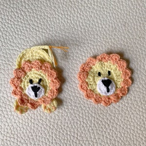 May include: Two crocheted lion appliques, one with a mane and paws, the other without. Both are yellow with peach-colored manes and black eyes.