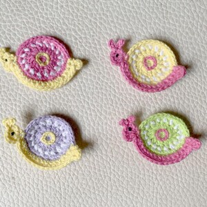 May include: Four crocheted snail appliques in different color combinations. Each snail has a pink body, yellow shell, and a white center with a smaller circle in the center. The snails are arranged in a square pattern on a light beige surface.
