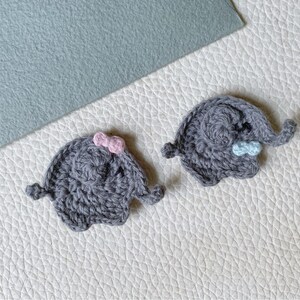 May include: Two handmade gray crochet elephant appliques. One has a pink bow, the other a blue bow. The appliques are on a white textured surface with a light blue felt square in the background. The appliques are small and detailed.