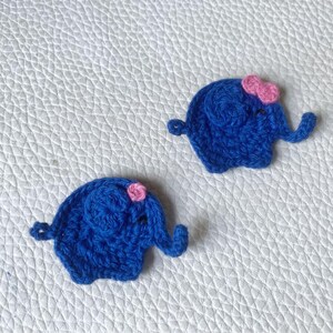 May include: Two small, dark blue crocheted elephant appliques, each featuring a tiny pink flower detail on the head. The elephants are crafted with visible yarn stitches, showing a rounded body, a trunk, and a small ear, with the text PUNT ART visible.