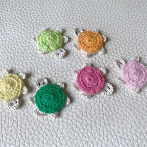 May include: A collection of seven colorful, crocheted turtle appliques. Each turtle features a spiral-stitched shell in shades of green, yellow, orange, pink, and white, with white crocheted flippers. The appliques are arranged on a textured, off-white surface.