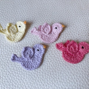 Könnte beinhalten: Vier gehäkelte Vogel-Applikationen in Pastellfarben: Gelb, Rosa, Lila und Fuchsia. Jeder Vogel hat ein kleines schwarzes Auge und einen orangefarbenen Schnabel. Die Vögel sind auf einer strukturierten weißen Oberfläche angeordnet.
