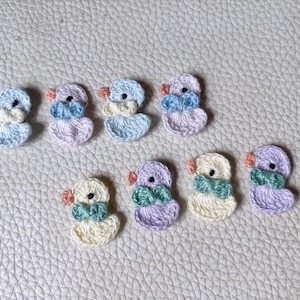 May include: A collection of handmade crochet ducklings in various pastel colors, including blue, pink, yellow, and purple. Each duckling features a contrasting bow tie and orange beak. The ducklings are arranged on a textured, off-white surface.