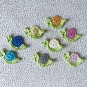 May include: A collection of handmade crochet snail appliques. Each snail features a green body and a shell in various colors, including blue, orange, pink, and white. The appliques are arranged on a textured white surface.