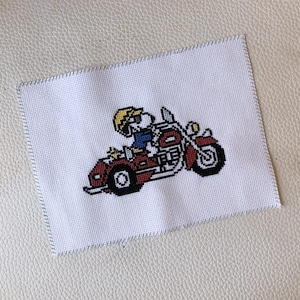 May include: Cross-stitch embroidery on white fabric featuring a cartoon character riding a red motorcycle with a sidecar. The character wears a yellow helmet and sunglasses. The motorcycle has black tires and chrome details.