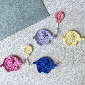 May include: Crocheted elephant appliques in pink, purple, yellow, and blue, with a balloon attached to two of them. The appliques are handmade and placed on a white textured surface. The appliques are small and detailed.