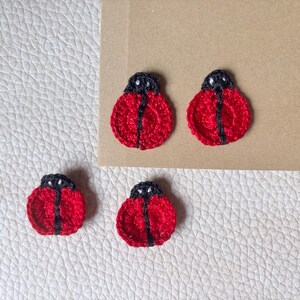 May include: Four small, circular ladybug appliques crafted from red and black yarn. Each piece features a vibrant red body with a central black stripe and a small black head section. The crochet texture is visible throughout the design.