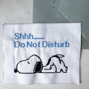 May include: A white cross-stitch with the text "Shhh... Do Not Disturb" in blue. The image features a black and white Snoopy character lying down. A gray envelope is in the background.