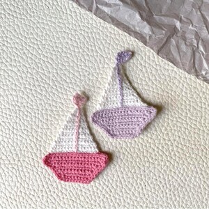 May include: Two handmade crochet sailboat appliques. One is pink and white, the other is purple and white. The appliques are on a textured, off-white surface. A piece of crumpled, gray paper is in the background.