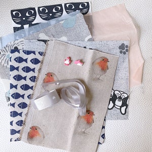 Puede incluir: Muestras de tela con diseños variados, incluyendo gatos, peces y petirrojos. Los colores son azul, gris y rosa, con una cinta blanca y pequeños elementos decorativos, perfectos para manualidades.