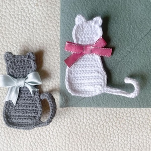 May include: Two crocheted cat ornaments. One is gray with a light blue bow, the other is white with a pink bow. Both have a tail and are sitting on a textured surface. The cat ornaments are handmade.