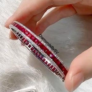 Pode incluir: Uma pulseira prateada com uma fileira de pedras preciosas vermelhas vibrantes e uma fileira de pedras preciosas claras menores. A pulseira é segurada por uma mão contra um fundo branco e fofo. O texto "DiamondsBySV" é visível.