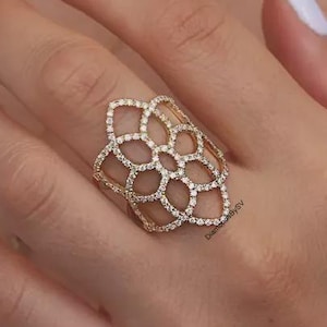 Op de afbeelding: Een goudkleurige ring met een ingewikkeld, opengewerkt bloem- of rasterontwerp. Het gehele oppervlak van het metalen frame is ingelegd met kleine, heldere, sprankelende steentjes. De ring bedekt een aanzienlijk deel van de vinger.