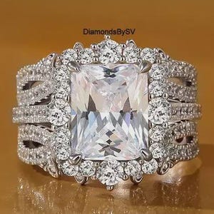 May include: A silver-toned ring featuring a large rectangular center stone surrounded by a halo of smaller round stones. The band is wide with multiple rows of pavé-set stones and intricate metalwork. The text DiamondsBySV is visible.