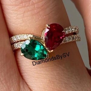 Puede incluir: Un anillo de oro con dos piedras preciosas en forma de pera, una verde esmeralda y otra roja rubí. El anillo está adornado con pequeñas piedras preciosas transparentes a lo largo de la banda. El texto "DiamondsBySV" es visible.