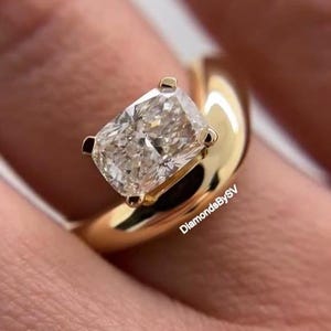 Op de afbeelding: Een ring met een radiant-geslepen diamant en een dikke, gepolijste gouden band. De diamant is gezet met vier klauwen en de ring heeft een modern, minimalistisch ontwerp. De ring wordt aan een vinger getoond.