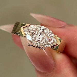 Puede incluir: Un anillo de oro con un diamante talla marquesa. El diamante está engastado horizontalmente, con un brillo visible. El anillo tiene un diseño moderno y minimalista. El texto "DiamondsBySV" es visible en el anillo.
