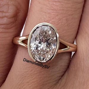 Puede incluir: Un anillo de oro con un diamante de talla ovalada. El diamante está engastado en un bisel y la banda tiene un diseño de vástago dividido. El texto "DiamondsBySV" es visible en el anillo.