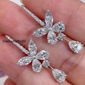 Könnte beinhalten: Ein Paar silberne Ohrhänger mit einem Schmetterlingsdesign mit birnenförmigen und runden, klaren Steinen. Die Ohrringe sind aus Metall und haben eine polierte Oberfläche.