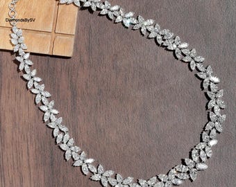 May include: A sparkling diamond necklace with a floral design. The necklace features numerous clear, brilliant-cut diamonds arranged in a delicate, leaf-like pattern. The necklace is displayed on a wooden surface.