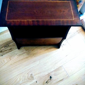 May include: A dark brown wooden side table with a rectangular top. The top has a lighter brown rectangular inlay with a thin gold border. The table has a curved apron and a lower shelf.