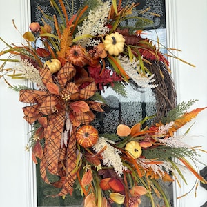 May include: A decorative wreath featuring a woven twig base adorned with artificial pumpkins in shades of orange, yellow, and cream. It includes brown plaid ribbon, dried grasses, green foliage, and autumn-colored leaves in red, orange, and brown tones.