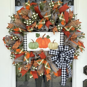 Könnte beinhalten: Ein herbstliches Kranz mit Bändern in Orange, Grün, Braun und Schwarz-Weiß-Mustern. Der Kranz hat ein zentrales Schild mit drei Kürbissen. Eine große schwarz-weiß karierte Schleife befindet sich unten.
