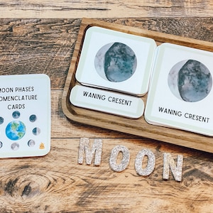 May include: A set of moon phase nomenclature cards with a wooden tray and the word "MOON" spelled out in glitter letters. The cards feature watercolor illustrations of the moon in different phases, including waning crescent.