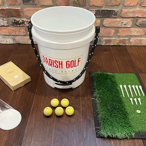May include: A white golf bucket with the text "Parish Golf Range Bucket" printed on the side. The bucket is on a wooden floor with a green golf mat, golf balls, and tees.