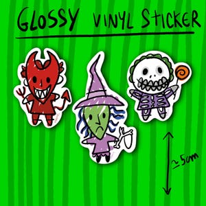 May include: Glossy vinyl stickers featuring three cartoon characters: a red devil, a green witch, and a skeleton-like figure holding a lollipop. The stickers are on a green striped background. The text "Glossy Vinyl Sticker" is at the top.