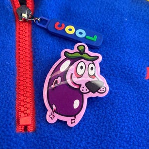 Peut inclure: Gros plan sur une veste polaire bleue avec une fermeture éclair rouge. Un autocollant de personnage de dessin animé, un chien-aubergine, est sur la veste. L'autocollant a un corps violet, des yeux blancs et un nez noir. Un tag bleu avec le mot "cool".