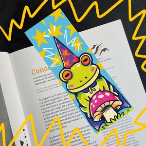 Könnte beinhalten: Ein farbenfroher Lesezeichen mit einem Cartoon-Frosch, der einen Partyhut trägt und einen Pilz hält. Der Lesezeichen hat einen blauen Hintergrund mit gelben Sternen und weißen Zickzacklinien. Der Frosch ist grün, mit orangen Augen und einem rosa Pilz.