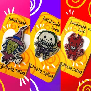 Puede incluir: Tres pines hechos a mano sobre tarjetas amarillas con el texto "handmade Love" y el nombre del artista. Los pines presentan personajes de dibujos animados: una bruja, un esqueleto y un diablo, cada uno con diseños y colores únicos. El fondo es una mezcla de rosa, morado y rojo.