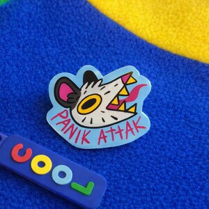 Peut inclure: Épinglette bleue avec un visage de monstre de dessin animé et le texte "PANIK ATTAK" en rouge. Un bracelet en caoutchouc bleu avec le mot "COOL" en lettres colorées est également visible.