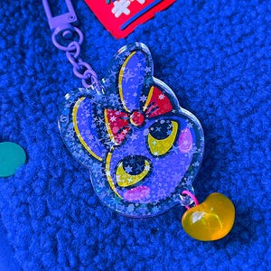 May include: A purple acrylic keyring featuring a stylised character face with large yellow eyes, a red bow, and a holographic star pattern. The keyring includes a purple lobster clasp and a small, translucent yellow heart-shaped charm attached.