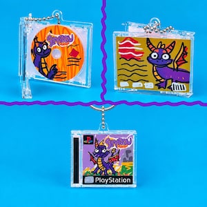 Puede incluir: Tres llaveros de estuche de CD en miniatura de plástico transparente con ilustraciones de Spyro the Dragon. Uno muestra un disco con patrones naranjas, otro una tarjeta dorada con un dragón morado y el tercero una portada con Spyro y PlayStation.