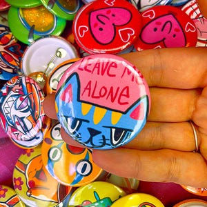 Op de afbeelding: Een ronde button met een illustratie van een blauwe kat met gele strepen op een roze achtergrond. De tekst "LEAVE ME ALONE" staat in het rood bovenaan. De button wordt omringd door een verzameling van diverse andere kleurrijke grafische pins.
